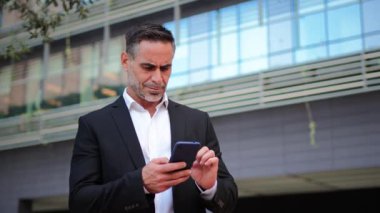 Deneyimli İşadamı: Olgun bir profesyonelin portresi cep telefonu ofis dışındaki iş e-postalarını kontrol ediyor, iş ve rahatlama arasında bir denge anı yakalıyor. Yüksek