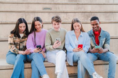 Merdivenlerde oturan, cep telefonlarını kullanarak sosyal medyada fotoğraf paylaşan bir grup çok ırklı genç arkadaş. Ergen öğrenciler bir toplantıda cep telefonu uygulaması ile birlikte oyun oynuyorlar.