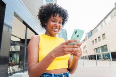 Afrikalı Amerikalı bir kız cep telefonuyla internete girip bir uygulama kullanarak gülümsüyor. Sosyal medyada cep telefonuyla fotoğraf paylaşan genç bir kız. Dışarıda akıllı telefon tutan genç bir kadın. Yüksek