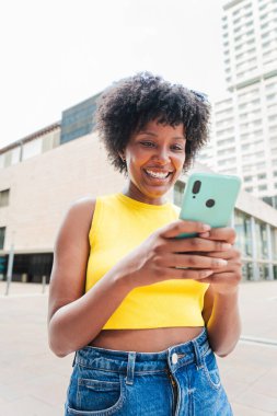 Dikey. Afrikalı Amerikalı bir kız cep telefonuyla internete girip bir uygulama kullanarak gülümsüyor. Sosyal medyada cep telefonuyla fotoğraf paylaşan genç bir kız. Elinde akıllı telefon tutan genç bir kadın. Yüksek