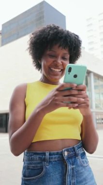 Dikey Afro-Amerikalı kız cep telefonuyla internete girip bir uygulama kullanarak gülümsüyor. Sosyal medyada cep telefonuyla fotoğraf paylaşan genç bir kız. Elinde akıllı telefon tutan genç bir kadın. Yüksek