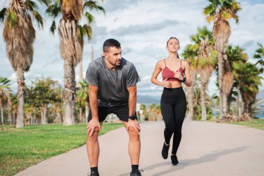 Erkek ve kadın dışarıda egzersiz yapıyor, palmiye çizgili bir yolda koşuyor, yoğun bir egzersizden sonra ara veriyor, spor faaliyetleri boyunca spor, dayanıklılık ve sağlıklı yaşam tarzı sergiliyor.