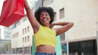 Bir Afro-Amerikalı genç bayan alışveriş merkezinden yeni kıyafetler aldıktan sonra sadece alışveriş çantalarını tutuyor. Genç müşteri kız kameraya bakarak gülümsüyor ve hediyelerini indirimde gösteriyor. Yüksek