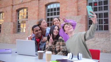 Bir grup neşeli öğrenci cep telefonuyla selfie çekerken dışarıda dizüstü bilgisayarla birlikte eğlenceli bir anı yakalıyorlar. Kampüste arkadaşlık, teknoloji ve öğrenci hayatı sergiliyorlar. Yüksek