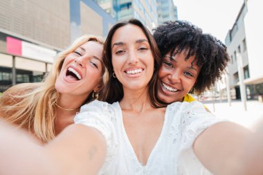Üç mutlu genç bayandan oluşan bir grup dışarıda selfie çekiyorlar, eğlenceli bir arkadaşlık, neşe ve kahkaha anı yakalıyorlar. Kızlar eğleniyor. Kadınlık, bağlanma, sosyal bağlantı ve kentsel yaşam tarzları
