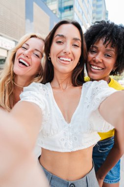 Dikey. Üç mutlu genç bayandan oluşan bir grup dışarıda selfie çekiyorlar, eğlenceli bir arkadaşlık, neşe ve kahkaha anı yakalıyorlar. Kızlar eğleniyor. Bağlanma, sosyal bağlantı ve kentsel yaşam tarzları