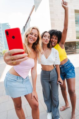 Dikey. Bir grup mutlu genç kadın dışarıda selfie çekiyor, gülümsüyor ve şehir ortamında birlikte eğleniyorlar, sosyal medyada paylaşmak için akıllı bir telefonla anıları yakalıyorlar.