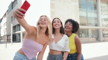 Üç mutlu genç bayandan oluşan bir grup dışarıda selfie çekiyor, birlikte bir şehir ortamında eğleniyor ve gülümsüyor, sosyal medya uygulamasında paylaşmak için kırmızı bir akıllı telefonla anıları yakalıyorlar.
