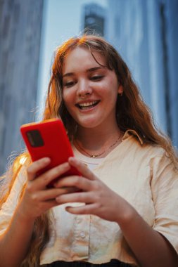 Şehirde geceleri akıllı telefon kullanan, sokak lambalarıyla aydınlatılmış dikey gülümseyen genç bir kadın. Akşamları teknoloji, iletişim, sosyal medya ya da şehir hayatı kavramları için mükemmel.