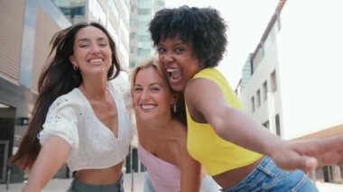 Üç mutlu, çok kültürlü genç kadın kameraya gülüyor ve el kol hareketi yapıyor. Açık havada, güneşli bir günde dostluk, eğlence ve neşe gösteriyorlar. Bir grup kadınsı kız