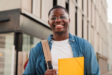 Gülümseyen Afrikalı Amerikalı öğrenci kameraya bakıyor, bir klasör tutuyor ve sırt çantası takıyor, modern bir şehir ortamında açık havada duruyor, parlak bir günde güven ve olumluluğu ifade ediyor. Yüksek