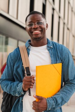 Dikey. Gülümseyen Afrikalı Amerikalı öğrenci kameraya bakıyor, bir klasör tutuyor ve sırt çantası takıyor, modern bir şehir ortamında açık havada duruyor, özgüven ve olumluluğu ifade ediyor. Yüksek