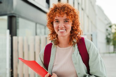 Kırmızı bir klasör tutarken ve sırt çantası takarken kameraya gülümseyen kızıl saçlı öğrenci, şehir ortamında dışarıda duruyor, kendine güveni ve pozitif bir tavrı parlak bir günde gösteriyor. Yüksek