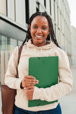 Dikey gülümseyen Afrikalı Amerikalı öğrenci elinde bir klasör tutarken kameraya bakıyor, şehir ortamında ayakta duruyor, güveni, eğitimi ve başarıyı dostça bir ifadeyle temsil ediyor.