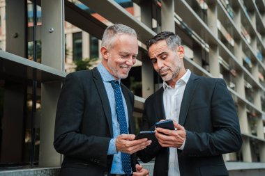 Takım elbiseli olgun iş adamları dışarıda duruyor, akıllı telefonları işle ilgili görevler için kullanıyor, bilgi paylaşıyor ve modern bir şehir ortamında iş stratejilerini tartışıyorlar. Profesyonel işbirliği.