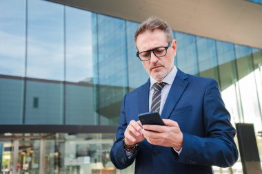 Modern bir ofis binasının önünde cep telefonu kullanan, iş ve iletişim odaklı, şehir merkezindeki akıllı telefon ve iş dünyasının profesyonel kullanımını gösteren resmi takım elbiseli bir iş adamı. Yüksek