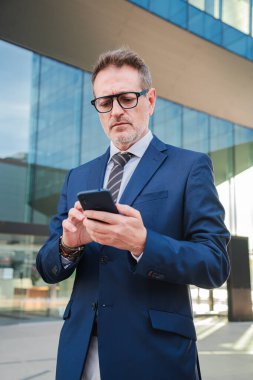 Dikey. Modern bir ofis binasının önünde cep telefonu kullanan resmi takım elbiseli, iş ve iletişim odaklı, şehir merkezinde profesyonel telefon ve iş kullanımını gözler önüne seren olgun bir işadamı.