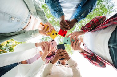Dışarıda cep telefonu kullanan bir grup arkadaş çemberin içinde dikiliyorlar. Canlı akıllı telefon renkleri, şehir ortamında modern teknolojiyi ve sosyal bağlantıyı gösteren gökyüzü ile zıtlık oluşturuyor. Yüksek