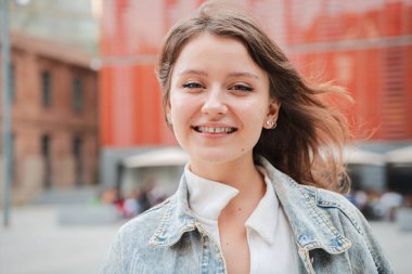 Genç bir kadın gülümsüyor ve dışarıdaki kameraya bakıyor, sıradan bir kot ceket giyiyor. Kendine güvenen ve mutlu bir ifade sergileyen genç bir kız doğal ışık ile kentsel bir çevrede kameraya bakıyor.