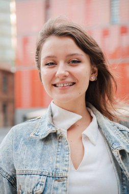 Dikey. Gülümseyen ve dışarıdaki kameraya bakan sıradan kot ceket giyen genç bir kadın. Kendine güvenen ve mutlu bir yüz ifadesini sergileyen genç bir kız doğal ışık alan bir şehirde kameraya bakıyor.