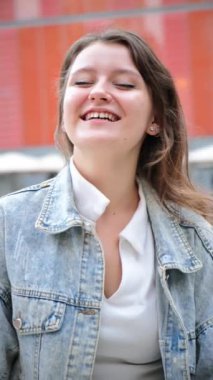 Dikey. Gülümseyen ve dışarıdaki kameraya bakan sıradan kot ceket giyen genç bir kadın. Kendine güvenen ve mutlu bir yüz ifadesini sergileyen genç bir kız doğal ışık alan bir şehirde kameraya bakıyor.