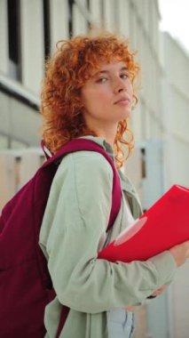 Dikey. Kızıl kıvırcık saçlı genç bir kız öğrenci modern bir kampüste açık havada dururken elinde parlak kırmızı bir dosya tutarak kameraya bakıyor. Düşünceli ifade, odaklanma ve akademik kararlılık