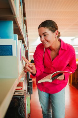 Kütüphanede kitap arayan genç bir kadın elinde bir kitap tutuyor ve rafları keşfediyor. Odaklanmış öğrenci veya okuyucu akademik bir kamu arşiv ortamında kitaplara göz atıyor