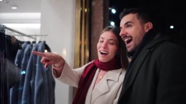 Gecenin tadını çıkaran genç bir çift, alışveriş poşetleri taşırken vitrin vitrinine heyecanlı bir şekilde bakıyor, şehir ışıklarıyla aydınlatılmış bir gece alışverişi sırasında neşeli bir anı paylaşıyor. Yüksek