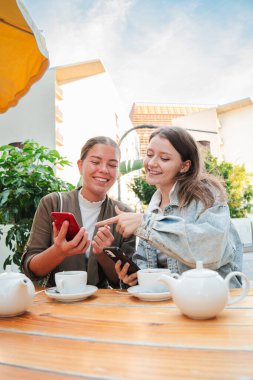 Dikey genç kadınlar birlikte çay içerken cep telefonlarını izleyip paylaşıyorlar, gülümsüyor ve dışarıda dinleniyorlar, sosyal medyayla iletişim kuruyorlar ve arkadaşça bir kahvehane terasında kaynaşıyorlar.