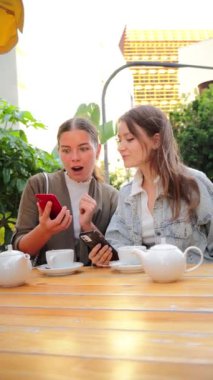 Dikey genç kadınlar birlikte çay içerken cep telefonlarını izleyip paylaşıyorlar, gülümsüyor ve dışarıda dinleniyorlar, sosyal medyayla iletişim kuruyorlar ve arkadaşça bir kahvehane terasında kaynaşıyorlar.