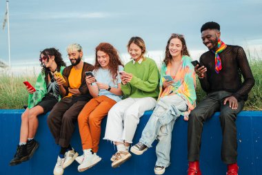 Çeşitli Arkadaş Grubu Akıllı Telefonları Birlikte Kullanırken Sosyal Medya 'da Neşeli Anı Paylaşır. Mavi Duvarda Sahil Gökyüzüne Karşı Oturan Genç İnsanlar İnternet 'te Cep telefonuyla geziyor. Yüksek