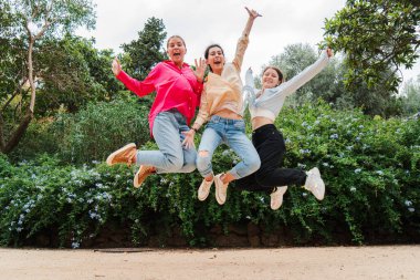 Heyecanlı genç kadınlar birlikte dışarı atlıyor, eğlenceli bir grup aktivitesinde neşe, arkadaşlık ve enerji ifade ediyorlar, yemyeşil bir park ortamında genç kızlar, kaygısız mutluluk anlarını yakalıyorlar. Yüksek