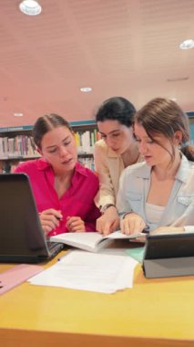 Dikey. Üniversite kütüphanesinde birlikte eğitim gören genç kadınlar, akademik kaynaklarla dolu raflarla çevrili masada kitaplarla, dizüstü bilgisayarlarla ödevler üzerinde işbirliği yapıyor ve fikirleri tartışıyorlar.