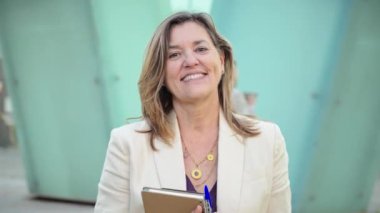 Kendine güveni tam bir iş kadını elinde bir defter ve kalem tutarken kameraya bakıyor, dışarıda duruyor. İş ortamında liderliği, profesyonelliği ve deneyimi temsil eden orta yaşlı bir kadın.