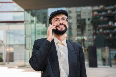 Hevesli, genç, sakallı girişimci akıllı telefonuyla anlamlı iş konuşmalarının keyfini çıkarırken şirket binasının önünde duruyor, kendine güveni ve yaklaşılabilir tavrını sergiliyor.