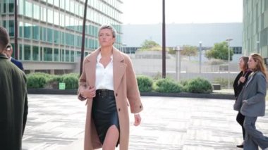 Kendine güvenen, kısa platin saçlı profesyonel iş kadını şık bej ceketli yoğun şirket meydanında kasten yürürken çeşitli meslektaşları modern ofis binasının önünden geçiyor.
