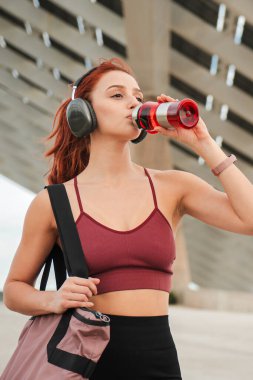 Spor yapan genç atletik kadın kulaklık takıyor ve açık hava kardiyo antrenmanından sonra şişeden izotonik su içiyor. Beden eğitimi rutininden sonra kadın susuzluk çekiyor. Yüksek