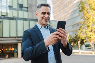 Açık havada cep telefonu kullanan olgun bir işadamı, takım elbiseli, gri saçlı ve sakallı profesyonel bir adam telefona bakıyor, şehir bölgesinde güvenle duruyor, teknoloji ve iletişimle ilgileniyor. Yüksek
