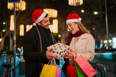 Sevgilisi, Noel Baba şapkası takmış ve bayram sezonunda etrafı şenlikli ışıklar ve alışveriş çantalarıyla çevrili sevgilisine Noel hediyesi veriyor. Yüksek