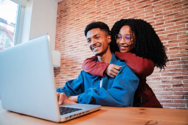 Mutlu Afro-Amerikan çift evde birlikte vakit geçiriyorlar, rahat bir masada dizüstü bilgisayar kullanıyorlar, sıcak ve rahat bir ortamda kucaklaşıp gülümsüyorlar. Gençler tatillerini internetten ayarlıyorlar