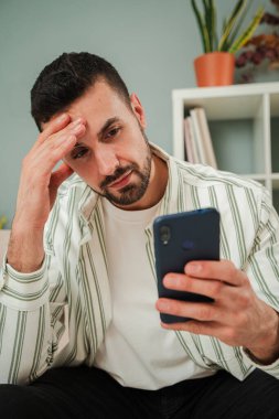 Endişeli genç adam elinde bir akıllı telefon tutuyor, kötü haberler okurken ya da bir sorunla yüzleşirken stresli ve hayal kırıklığına uğramış hissediyor, sıradan bir ortamda evde oturuyor, endişe ve düşünce gösterisi yapıyor. Yüksek