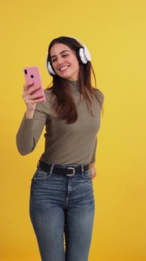 Dikey. Kulaklıklı ve akıllı telefonlu müzikten hoşlanan mutlu genç bir kadın, canlı sarı bir arka plana karşı canlı bir uygulamadan müzik eşliğinde dans ederek, neşe, eğlence ve modern dijital yaşam tarzını ifade ediyor.