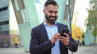 Kendine güvenen, olgun bir iş adamı, modern bir şehirde cep telefonu kullanıyor, resmi kıyafetler giyiyor, gülümsüyor ve üretken kalıyor, profesyonel başarıyı, iletişimi ve bağlantıyı gösteriyor.