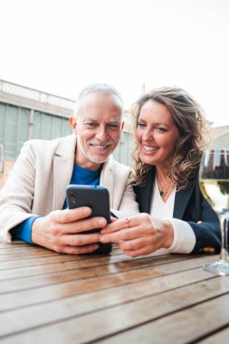 Dikey. Gülümseyen olgun bir çift, rahat bir bar terasında şaraptan keyif alıyor, akıllı telefon kullanıyor, mutlu anları paylaşıyor, bağ kuruyor ve masada beyaz şarap kadehleriyle dinleniyor. Orta yaşlı insanlar