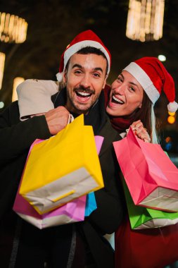 Dikey neşeli genç çift Noel şapkaları takıyor ve ellerinde renkli alışveriş çantaları tutuyorlar, gece vakti bayram alışverişinin tadını çıkarıyorlar. Parlak dekoratif kostüm ışıkları ve dışarıda neşeli ifadeler var.