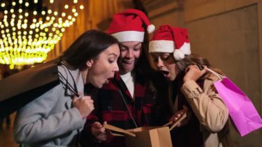 Noel şapkası ve kışlık giysiler giyen üç heyecanlı kadın, bayram ışıkları altında bir alışveriş çantasının içine hevesle bakıyor, gece kutlamalarında neşe ve sürprizleri paylaşıyor.