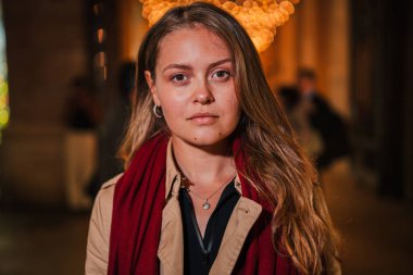 Uzun sarı saçlı, kırmızı eşarplı, ciddi bir genç kadın, gece festival ışıklarının altında dikiliyor, odaklanmış bir ifadeyle kameraya bakıyor, düşünceli tavırlı bir kadın önde duruyor.