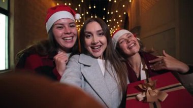 Noel Baba şapkası takan üç genç kadın gece bir video konuşmasında el sallıyor, neşeli ışıklarla aydınlatılıyor, Noel neşesini ve arkadaşlığını sıcak bir Noel bayramında kutluyorlar. Yüksek