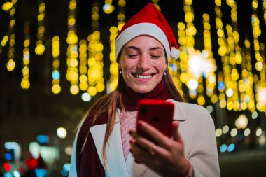 Noel şapkası takan gülümseyen genç bir kadın cep telefonunu kullanıyor, neşeli ışıklarla çevrili, büyülü bir Noel kutlamasında bayram sezonunun keyfini çıkarıyor.