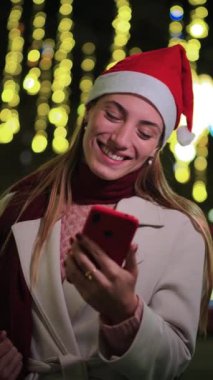 Dikey. Noel şapkası takan gülümseyen genç bir kadın cep telefonunu kullanıyor, neşeli ışıklarla çevrili, büyülü bir Noel kutlamasında bayram sezonunun keyfini çıkarıyor. Yüksek